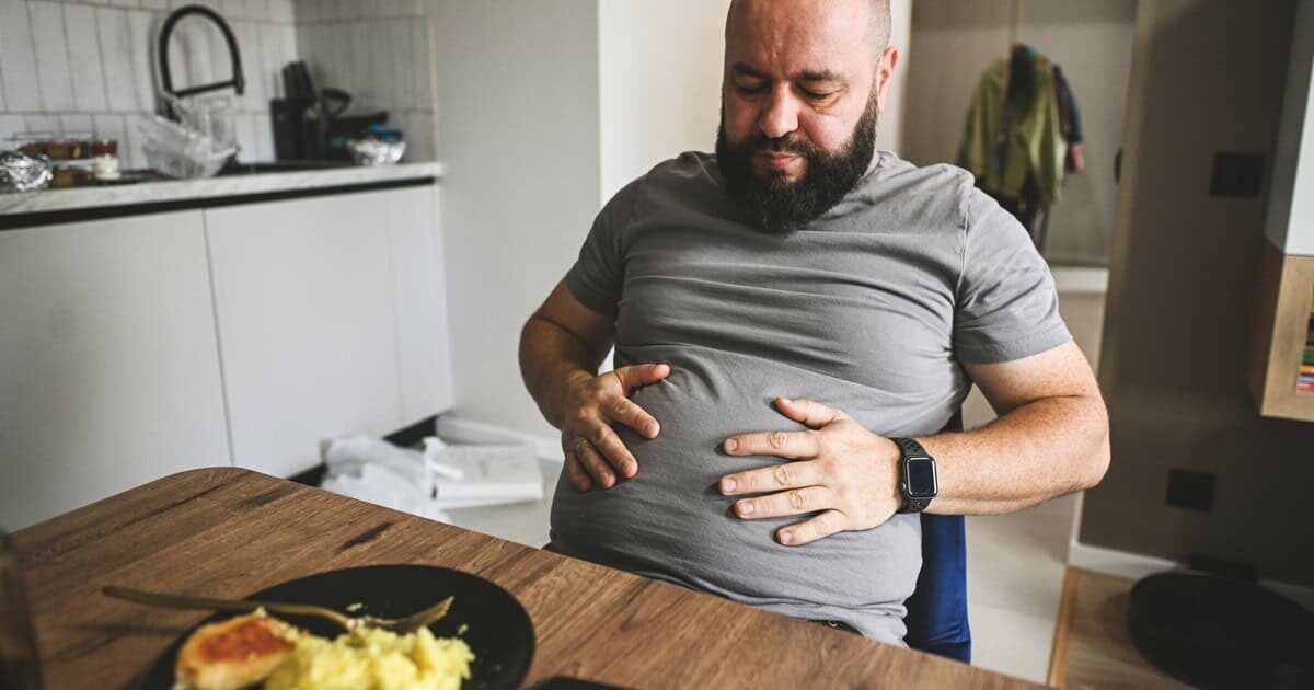 Muž v strednom veku sedí po obede pri stole a drží si rukami plné, nafúknuté brucho